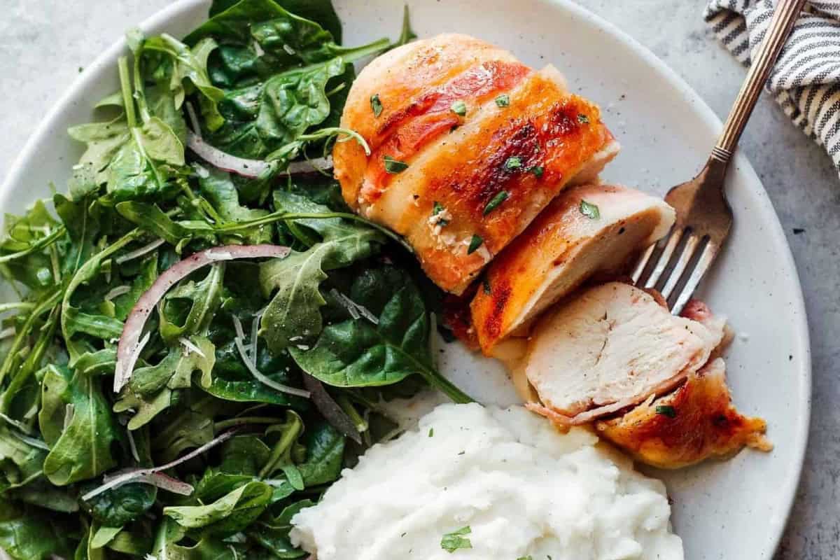 sliced bacon wrapped chicken next to salad and mashed potatoes.