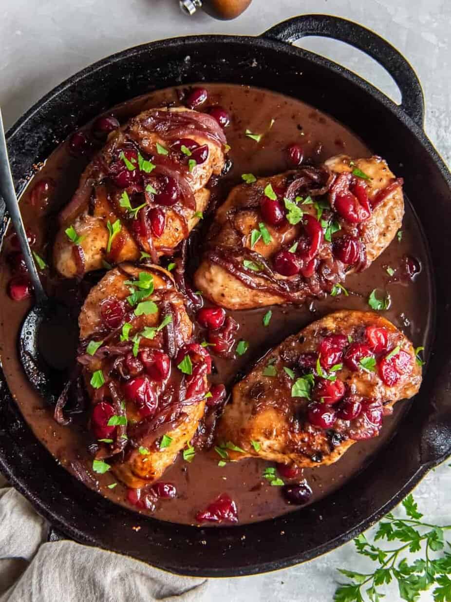 overhead view of 4 cranberry chicken breasts in a cast iron skillet with a spoon.