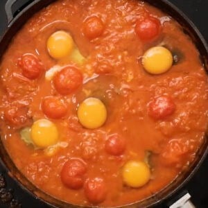 five raw eggs cracked into shakshua sauce in a pan.