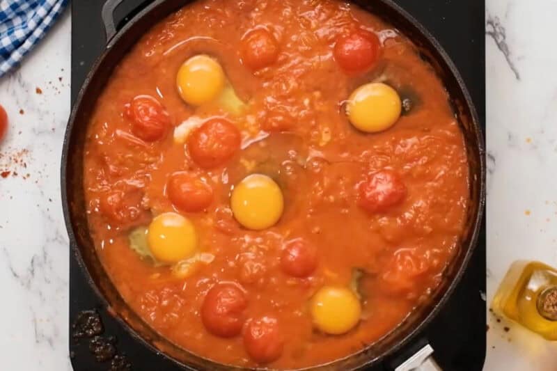 five raw eggs cracked into shakshua sauce in a pan.