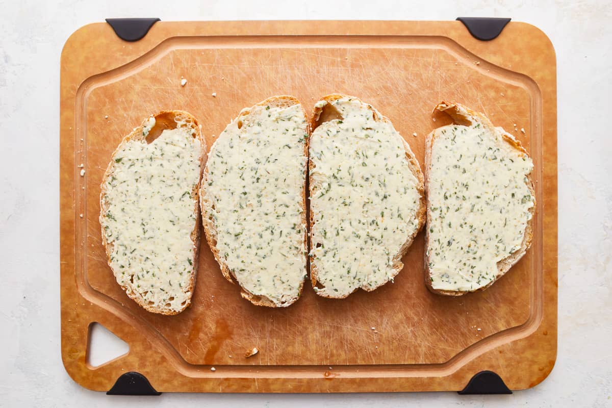 Four slices of sourdough bread topped with herb butter, on a wooden cutting board.