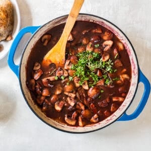 A pot of stew with mushrooms, tomatoes, and a wooden spoon.