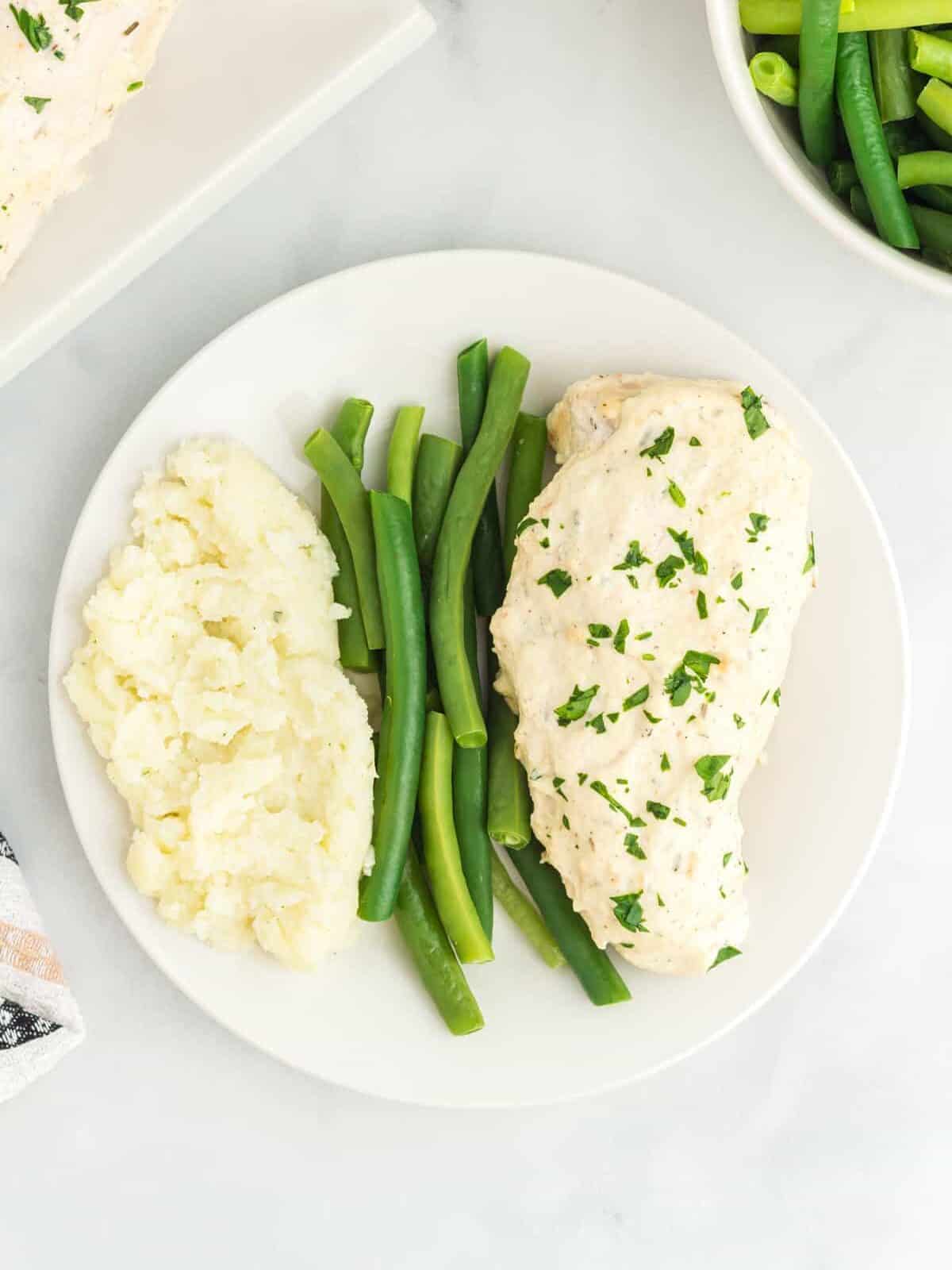 overhead view of a creamy herb chicken breast on a white plate with green beans and mashed potatoes.