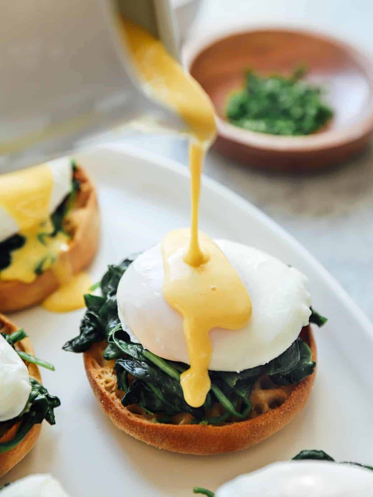 three-quarters view of a spoon pouring hollandaise sauce over eggs florentine benedict on a white serving tray.