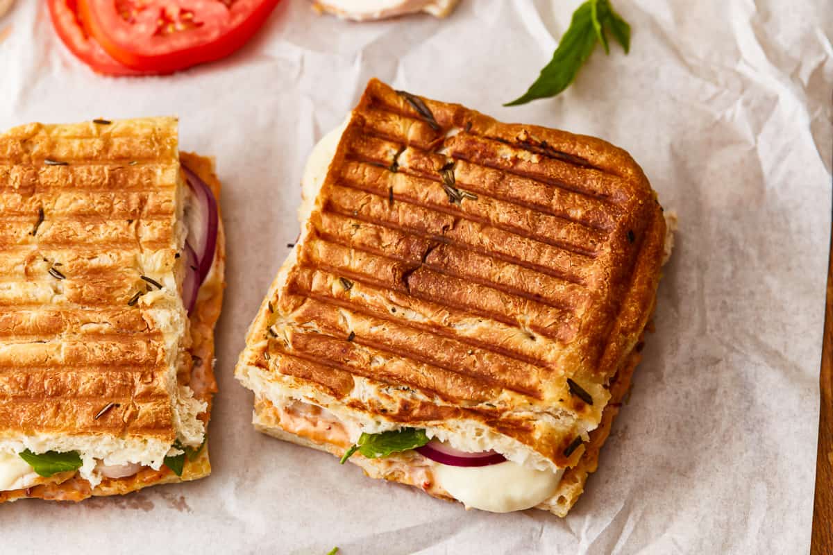 Two grilled sandwiches with tomatoes and onions on a piece of paper.