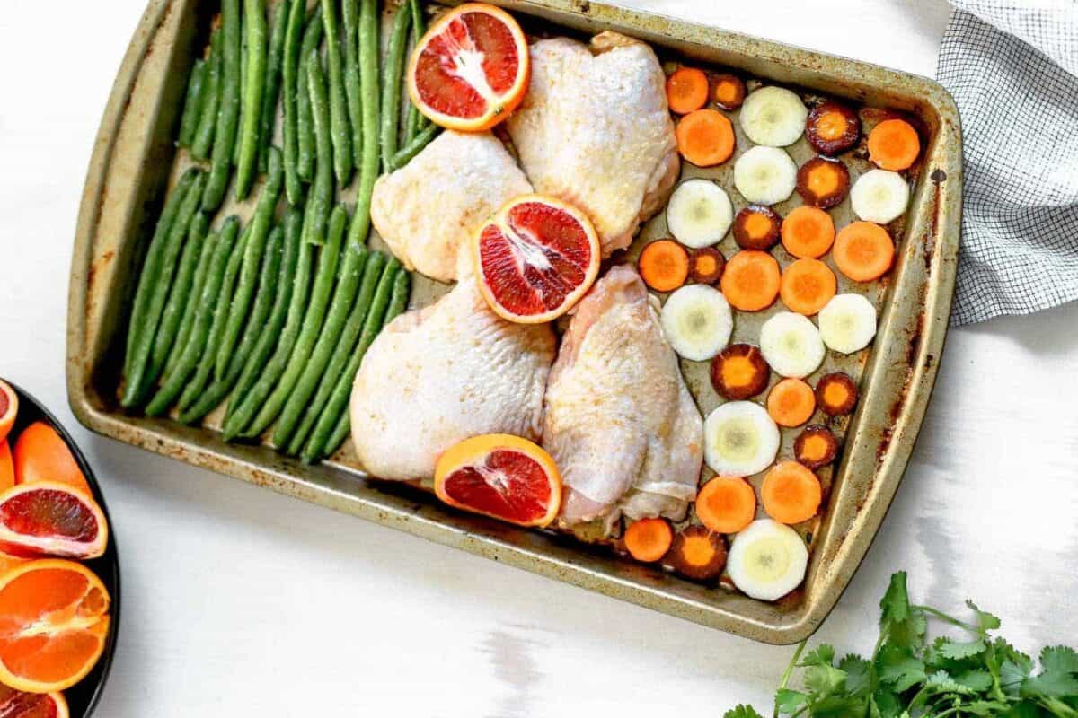 chicken thighs, blood oranges, sliced carrots, and stalks of asparagus spread out on a sheet pan.