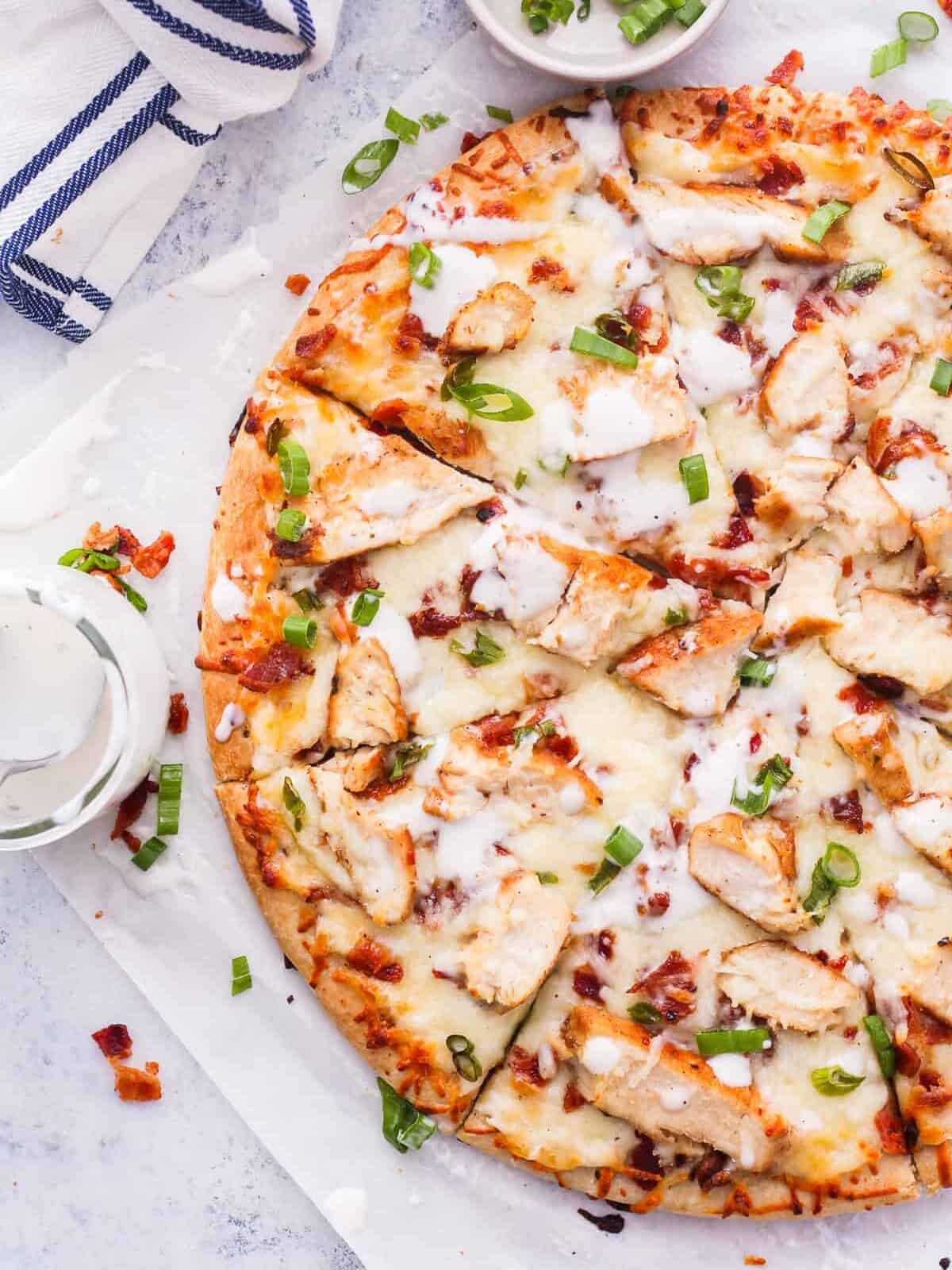 A chicken and cheese pizza on a white plate, topped with bacon and ranch dressing.