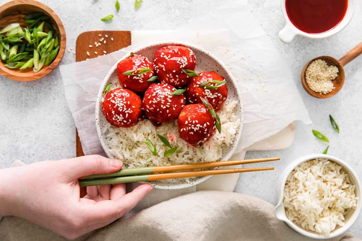 sweet and sour chicken meatballs with rice on a white plate with a hand holding chopsticks