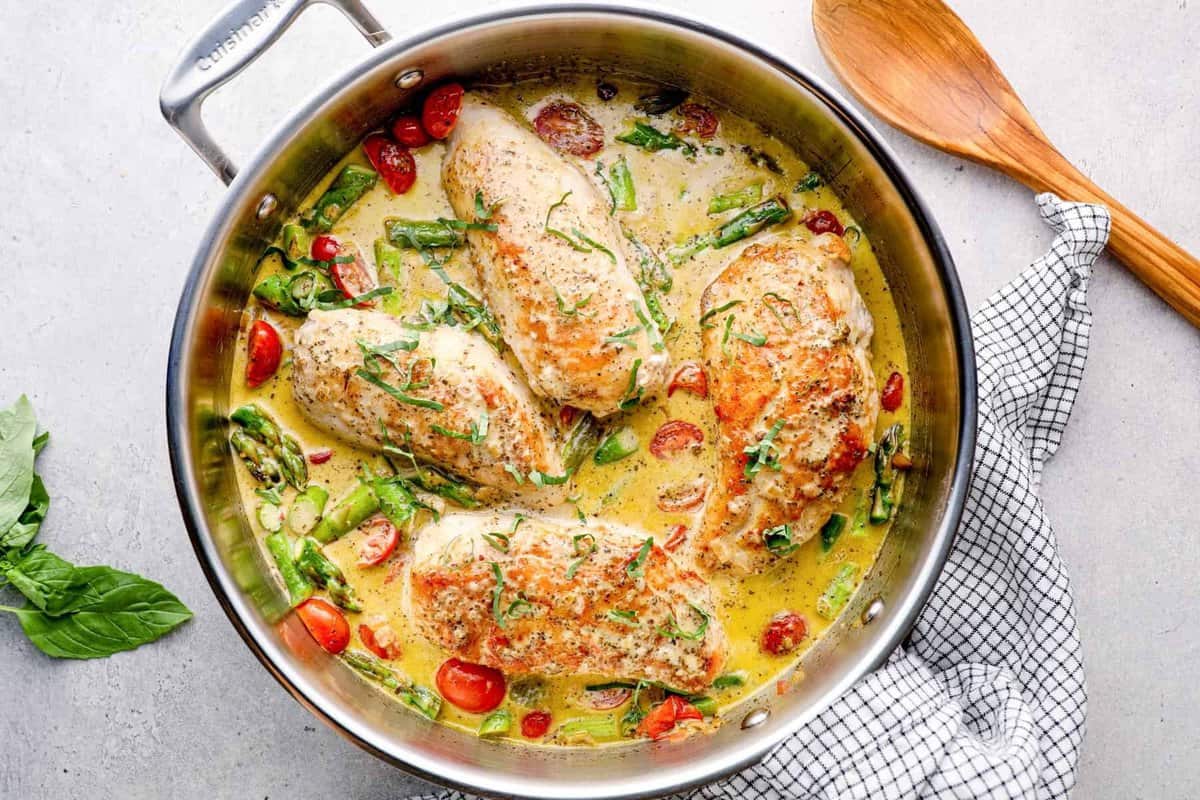 overhead image of chicken breasts in a skillet with pesto cream sauce, tomatoes, and asparagus