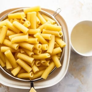 cooked rigatoni in a strainer set over a bowl next to a mug of pasta cooking water.