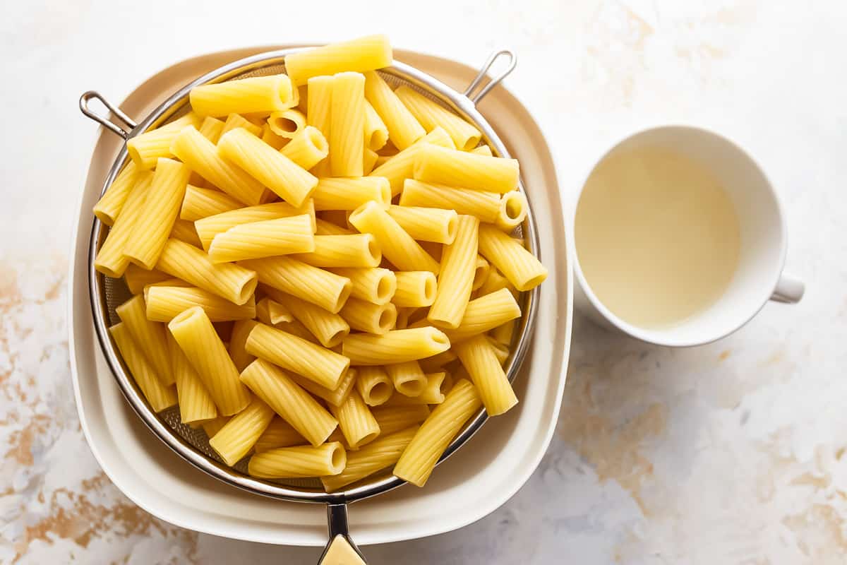 cooked rigatoni in a strainer set over a bowl next to a mug of pasta cooking water.