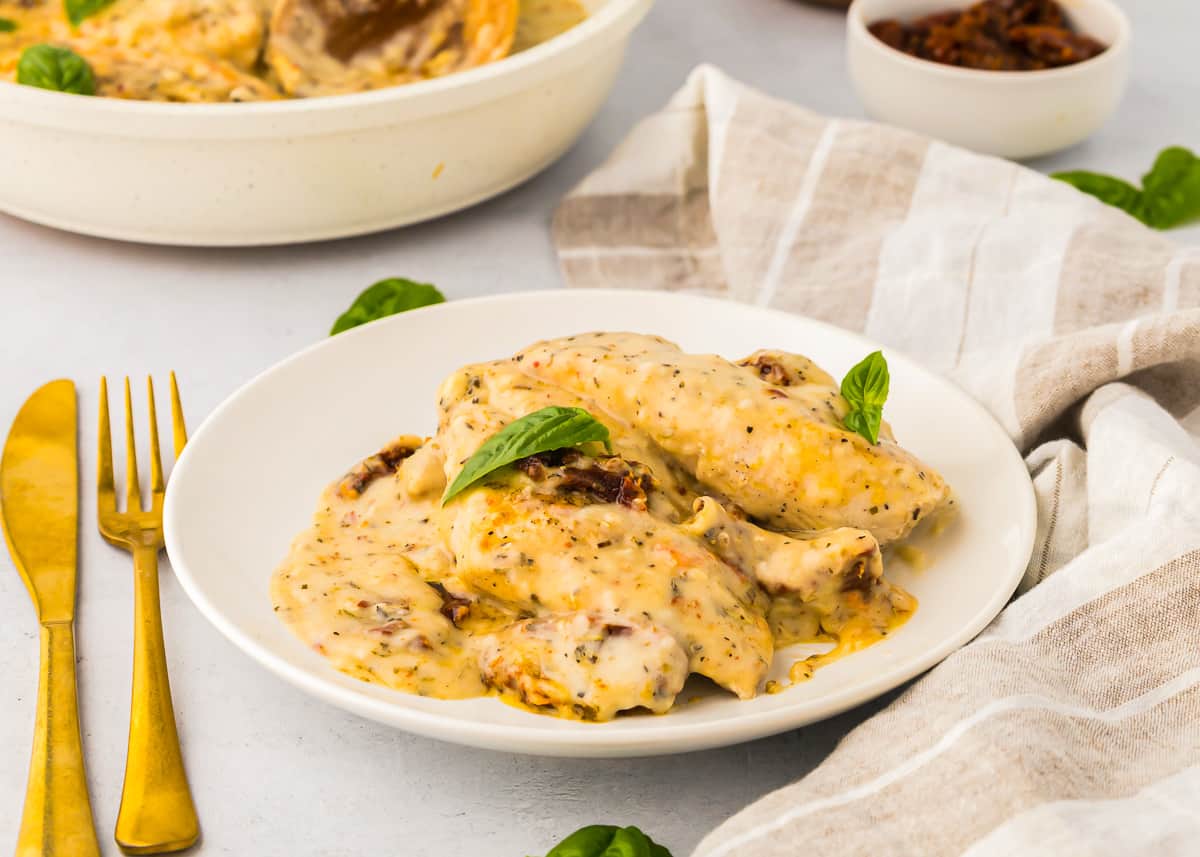 side view of a serving of marry me chicken on a white plate topped with fresh basil leaves.