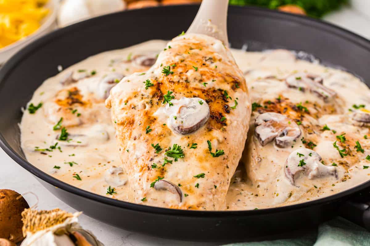 lifting a chicken breast from a pan of creamy chicken with a spoon.