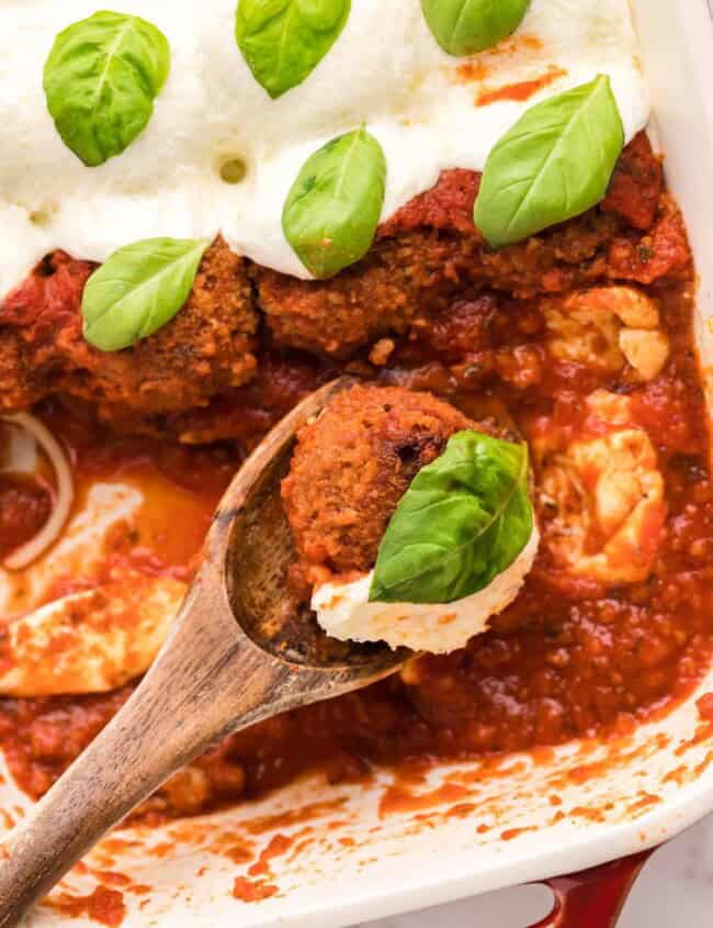 wood spoon held over a baking dish filled with chicken parmesan meatballs with one meatball on the spoon