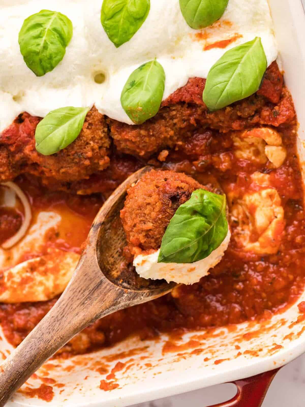 wood spoon held over a baking dish filled with chicken parmesan meatballs with one meatball on the spoon
