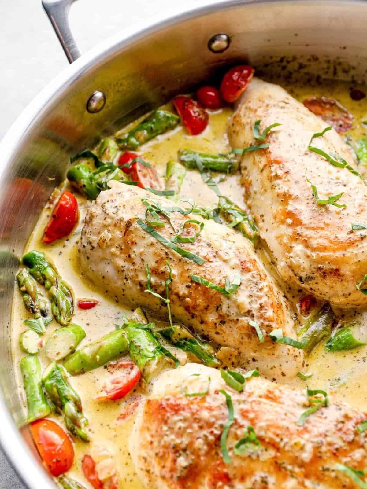 chicken breasts in a skillet with pesto cream sauce, tomatoes, and asparagus