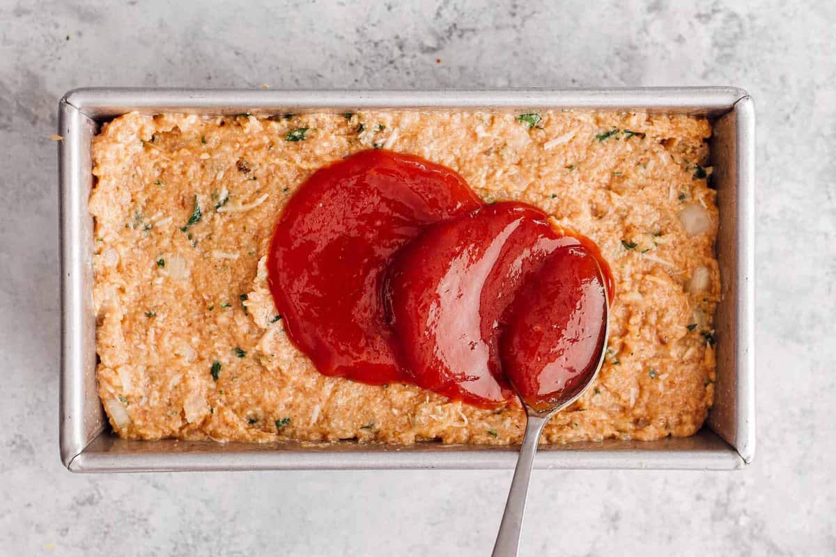 Spreading ketchup glaze on a chicken meatloaf in a loaf pan.