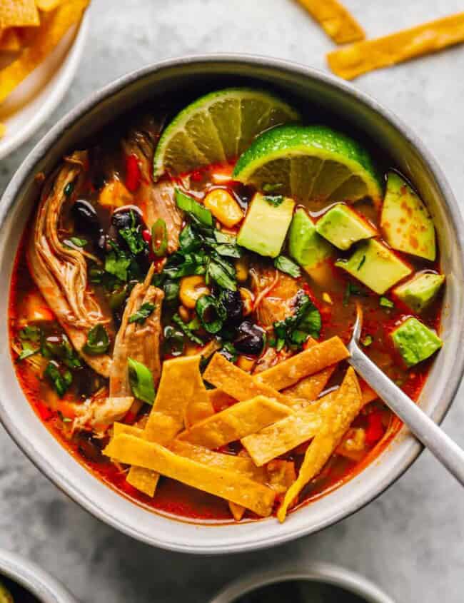 a bowl of authentic chicken tortilla soup, with chicken, tortilla strips, limes, avocado, and more