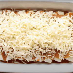 Enchiladas topped with sauce and shredded cheese before being baked.