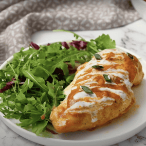 A stuffed buffalo chicken breast drizzled with ranch, on a plate with salad greens.