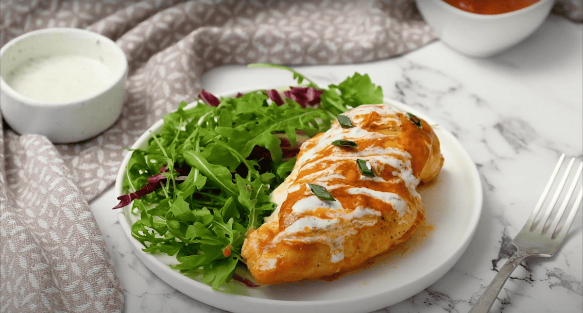 A stuffed buffalo chicken breast drizzled with ranch, on a plate with salad greens.