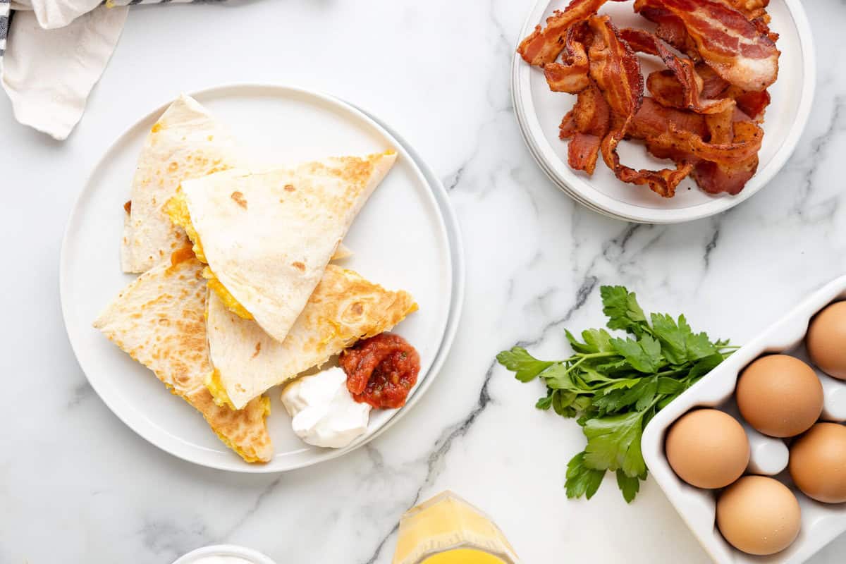 A plate of breakfast quesadillas, next to eggs and bacon on a countertop.