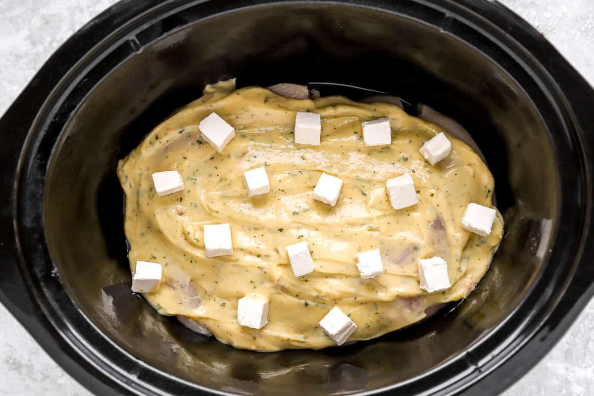 raw chicken breasts topped with ranch sauce and cubes of cream cheese in a crockpot.