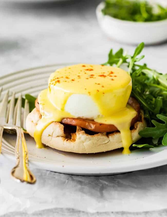 eggs benedict on a white plate with arugula and a fork.