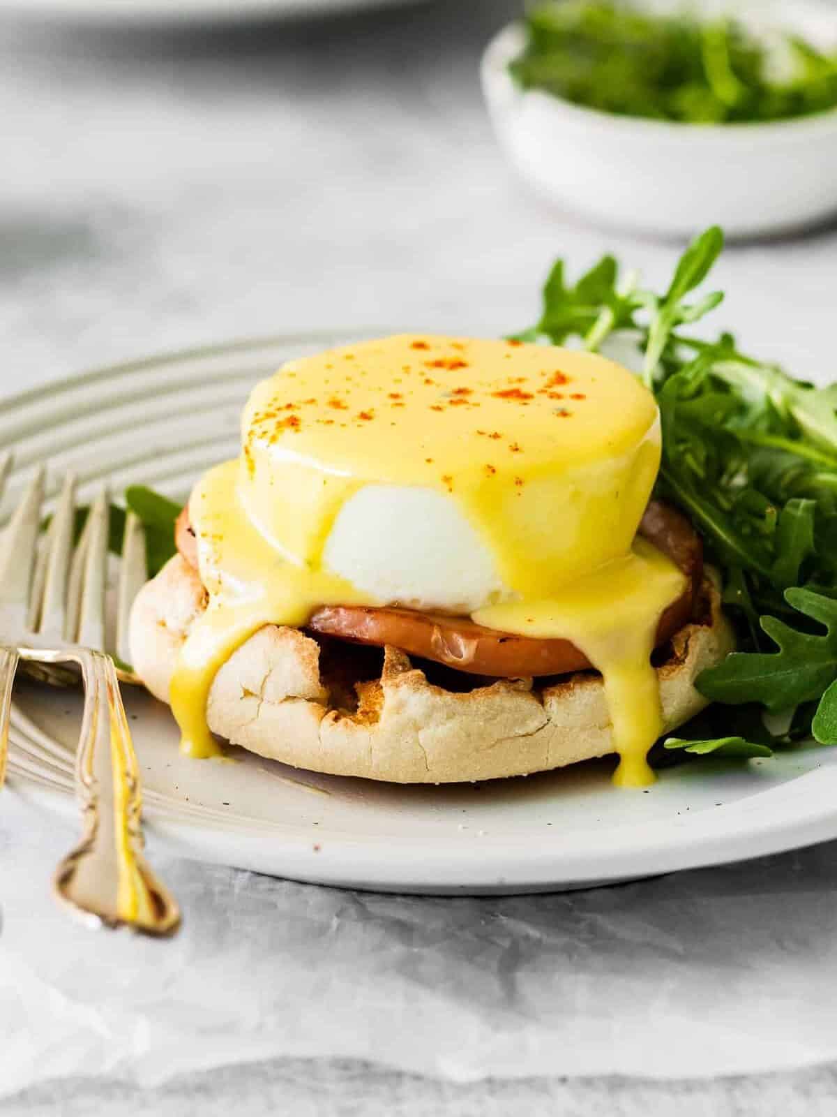 Eggs Benedict topped with hollandaise sauce on a white plate with arugula and a fork.
