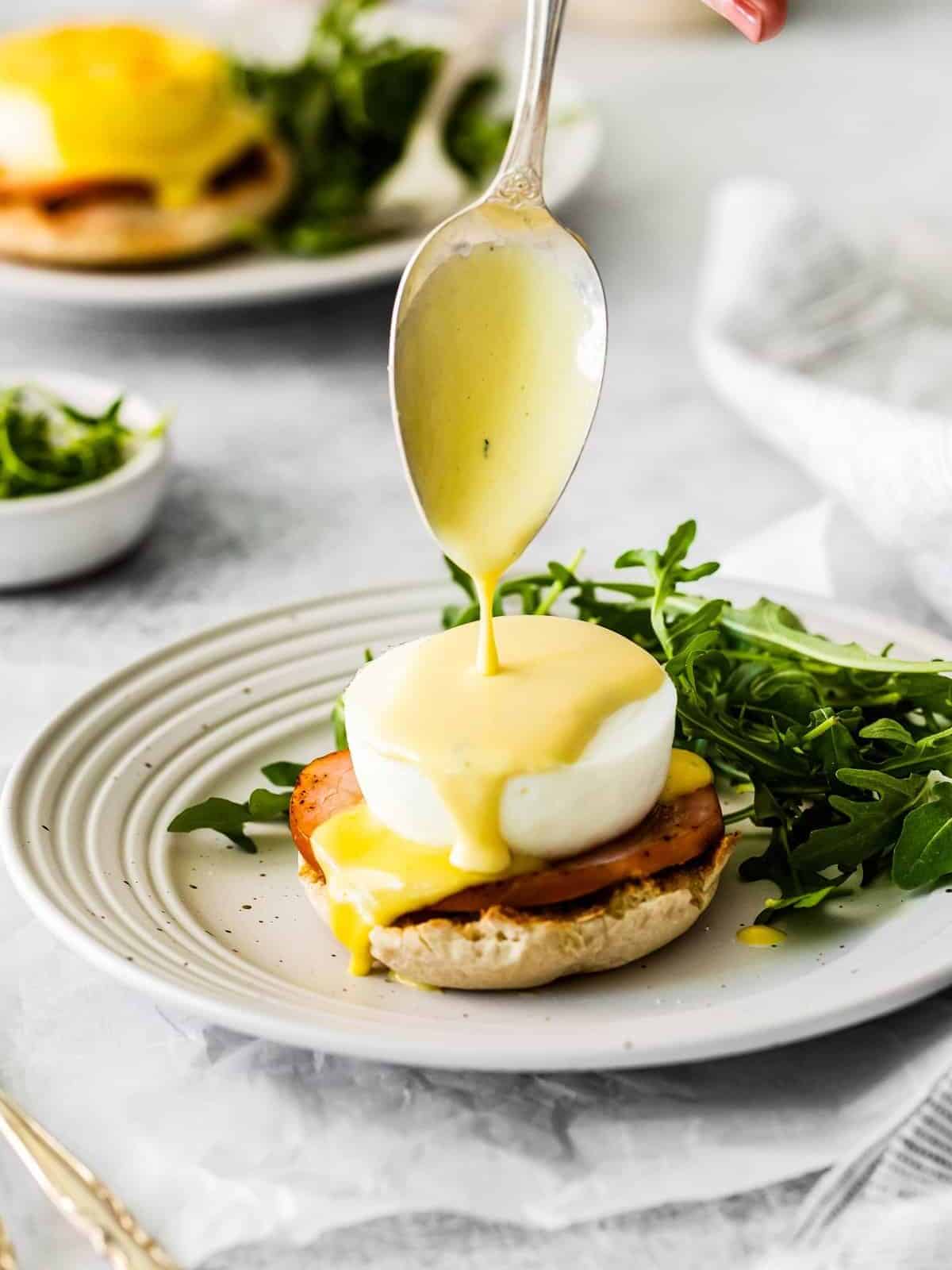 Hollandaise sauce drizzling off a spoon, on top of Eggs Benedict.