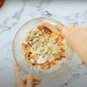 Glass mixing bowl filled with buffalo sauce, shredded chicken, cream cheese, and blue cheese.