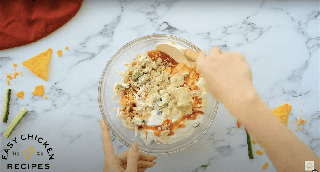 Glass mixing bowl filled with buffalo sauce, shredded chicken, cream cheese, and blue cheese.