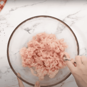 Crumbling raw ground chicken in a glass mixing bowl.