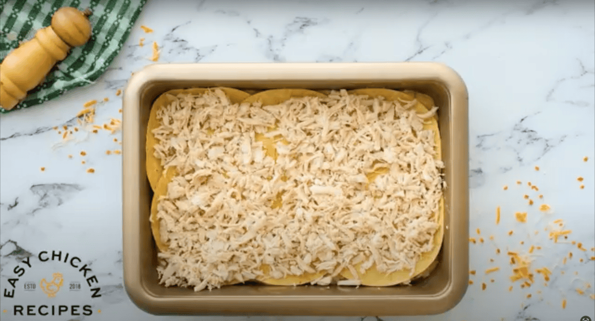 Shredded rotisserie chicken spread across a layer of tortillas in a baking dish.