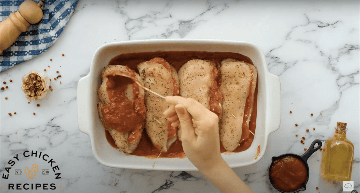 Spreading marinara on top of 4 stuffed chicken breasts in a baking dish.