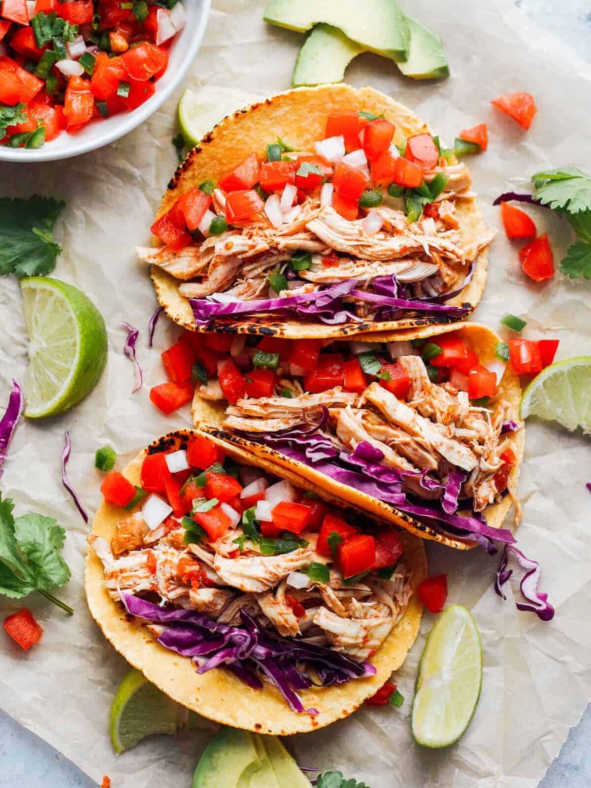 3 Instant Pot chicken tacos topped with purple cabbage and Pico de Gallo; with other fresh ingredients scattered around them.