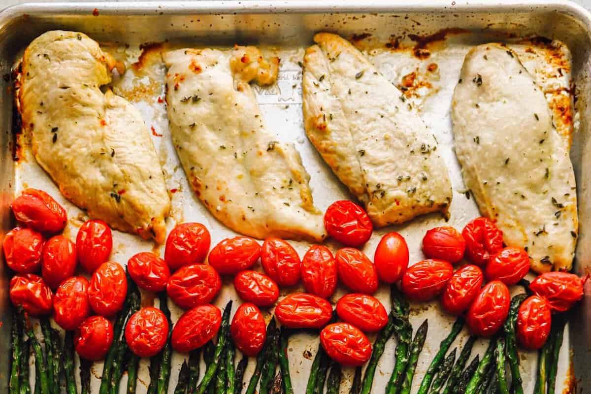 Garlic herb chicken breast, tomatoes, and asparagus arranged on a sheet pan.