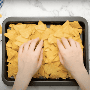 Hands spreading tortilla chips onto a baking tray.