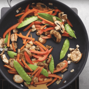 Carrots, red bell peppers, snow peas, and mushrooms cooking in a pan.