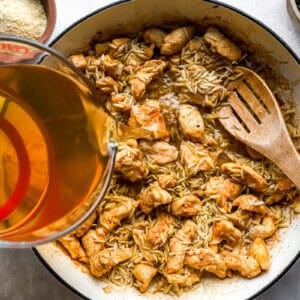 pouring broth over chicken pieces and orzo in a pan.