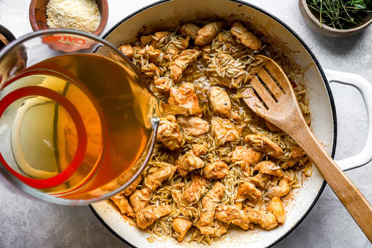 pouring broth over chicken pieces and orzo in a pan.