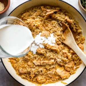 pouring cream over chicken and orzo in a pan.