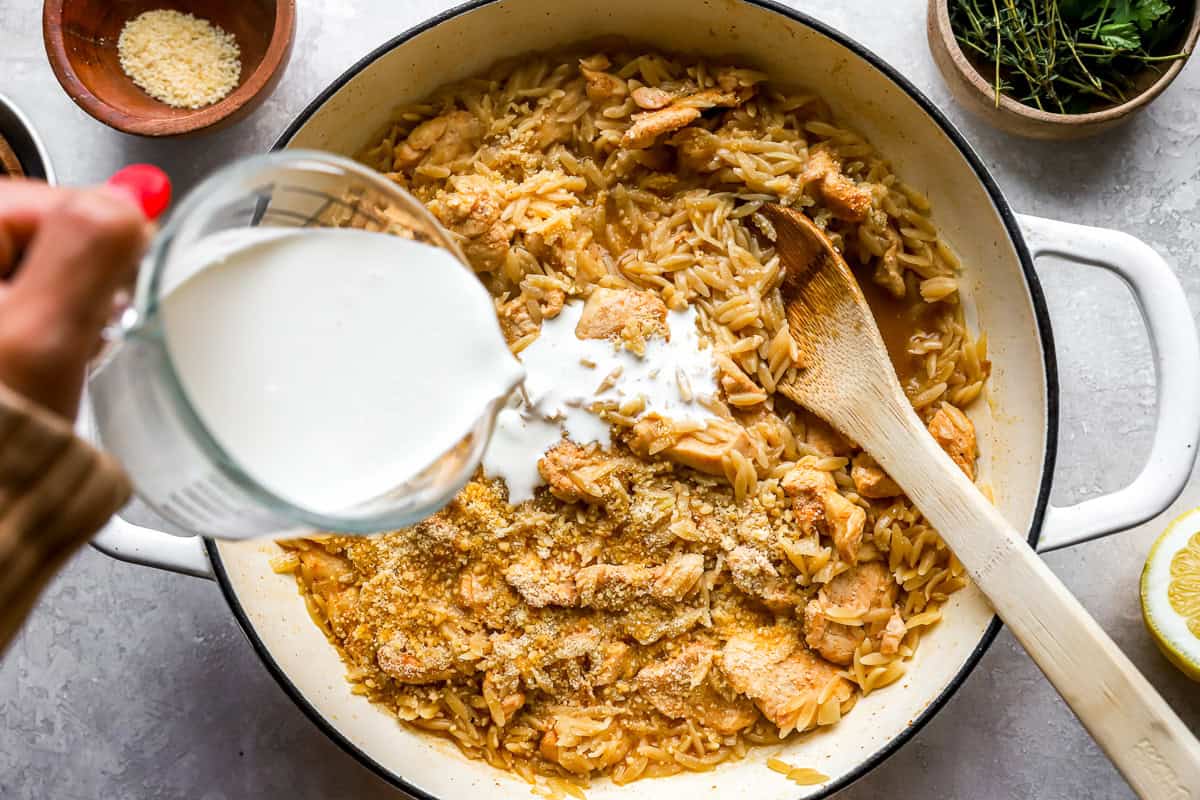 pouring cream over chicken and orzo in a pan.