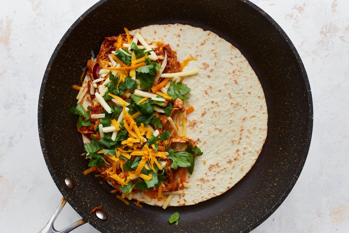 bbq chicken, cilantro, and cheese on half of a flour tortilla in a pan.