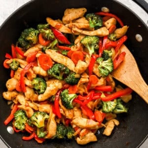 Pieces of chicken, broccoli, carrots, and bell peppers cooking in a skillet.