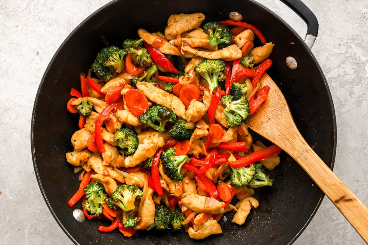 Pieces of chicken, broccoli, carrots, and bell peppers cooking in a skillet.