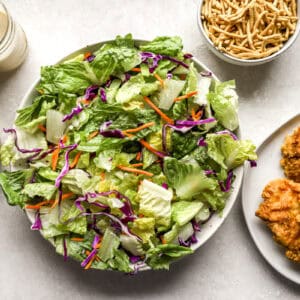 Bowl of salad greens next to a plate of crispy chicken, a jar of dressing, and a bowl of crispy noodles.