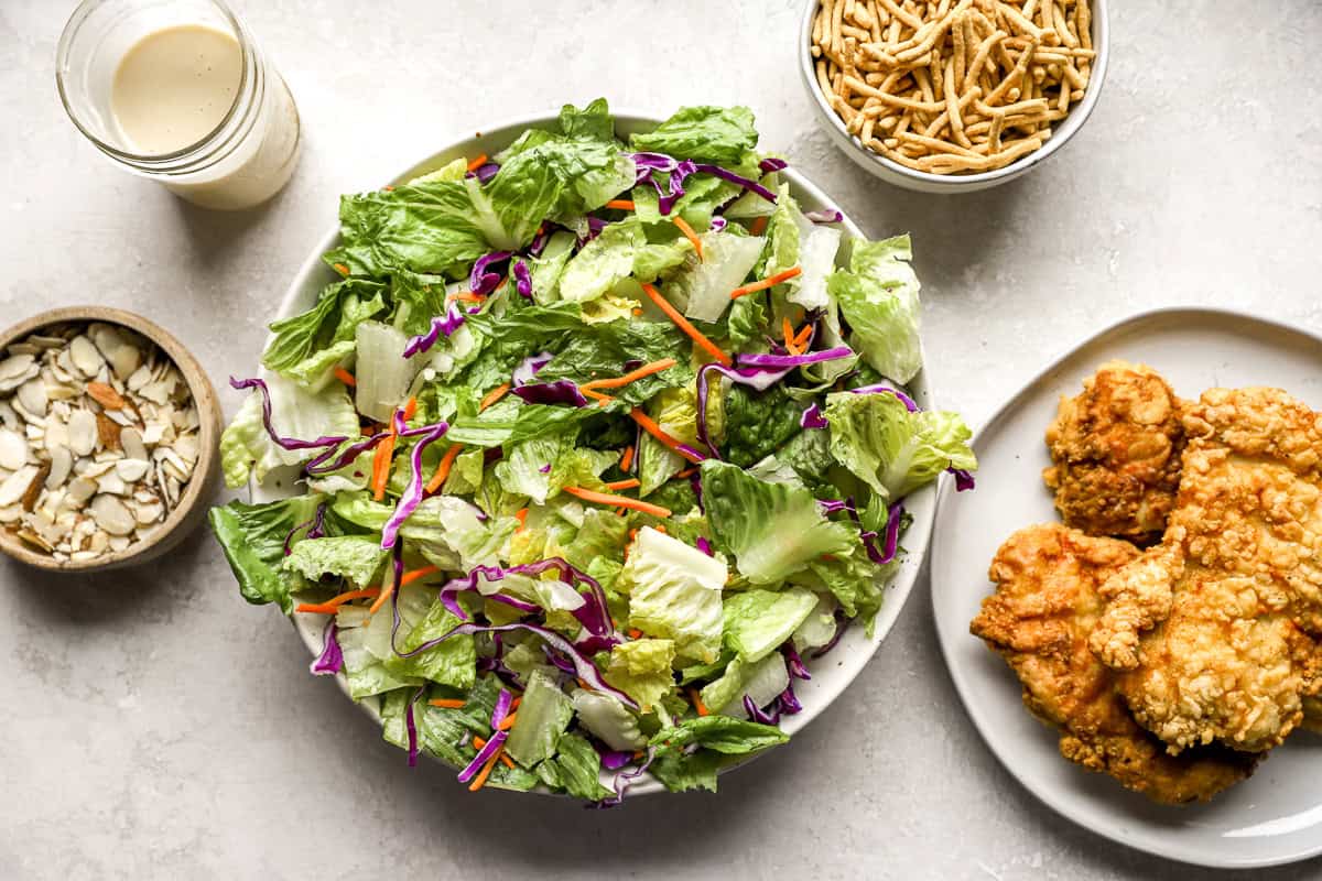 Bowl of salad greens next to a plate of crispy chicken, a jar of dressing, and a bowl of crispy noodles.