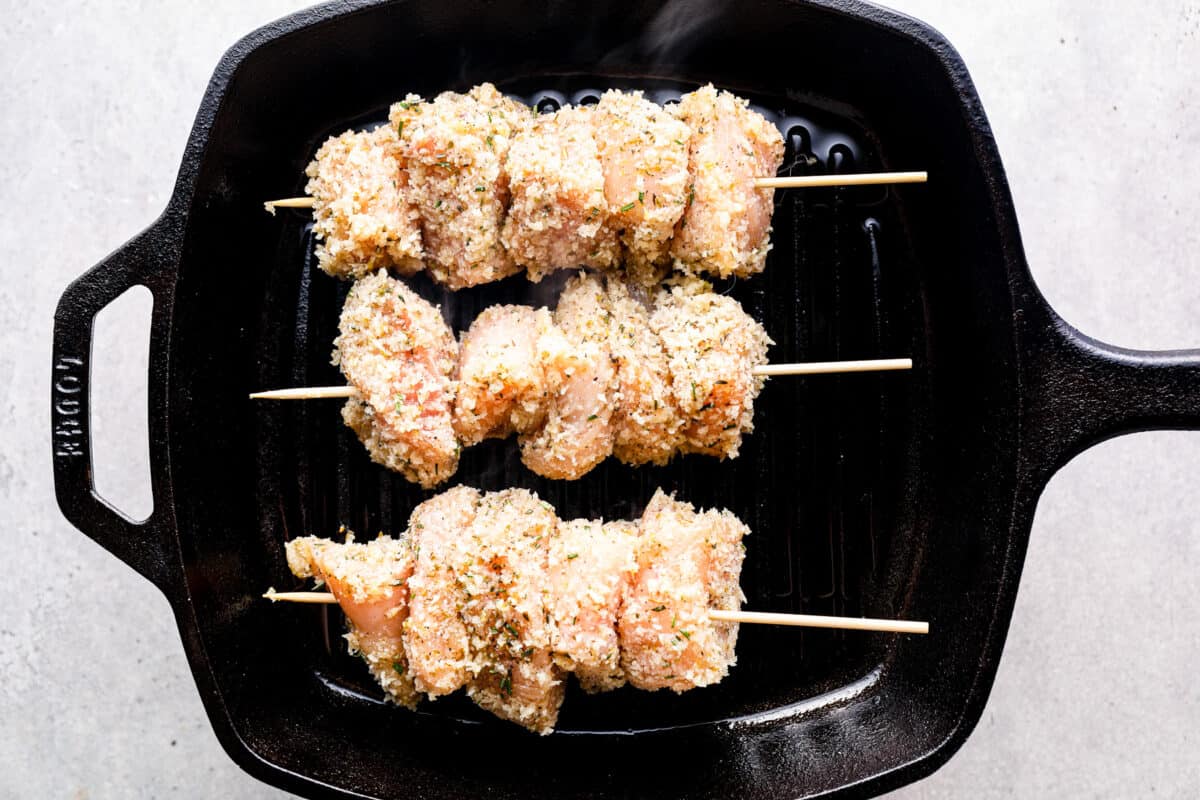 Breadcrumb-coated pieces of chicken on skewers, lined up on a grill pan.