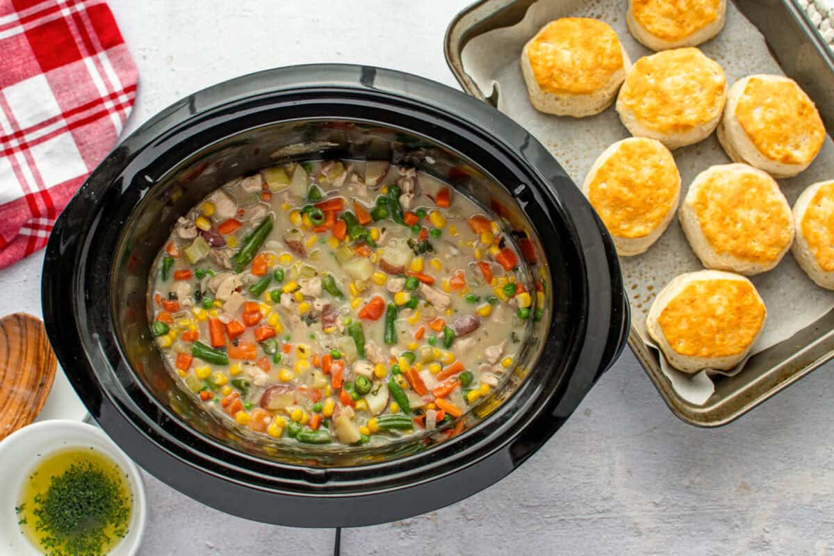 A slow cooker filled with chicken and veggie filling, next to a tray of biscuits.